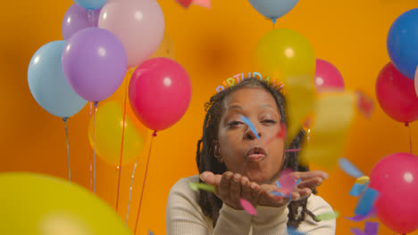 Studio-Portrait-Of-Woman-Wearing-Birthday-Headband-Celebrating-Blowing-Paper-Party-Confetti-To-Camera